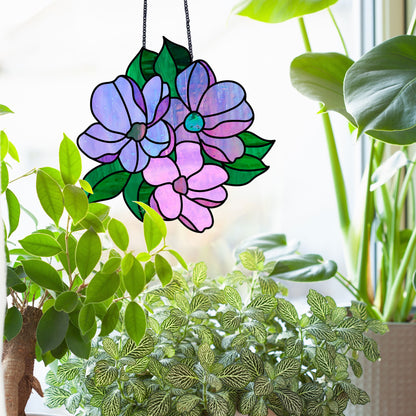 The Bundle of Flowers Stained Glass Pattern suncatcher, featuring purple blooms and green leaves, gracefully hangs in front of a window. Surrounded by lush indoor plants, it transforms natural light into a vibrant display.