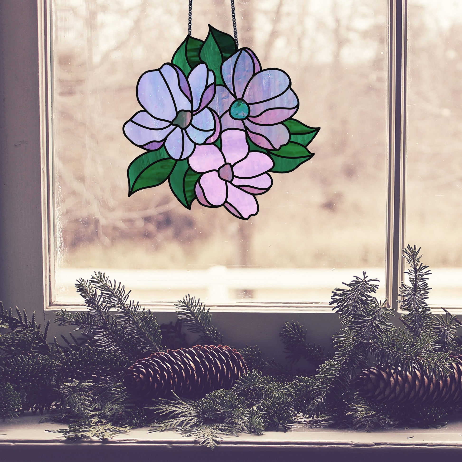 A stained glass suncatcher using the Bundle of Flowers Stained Glass Pattern in purple and pink hangs at the window. On the sill, pine branches and cones are artfully arranged against a blurred outdoor scene for a serene backdrop.