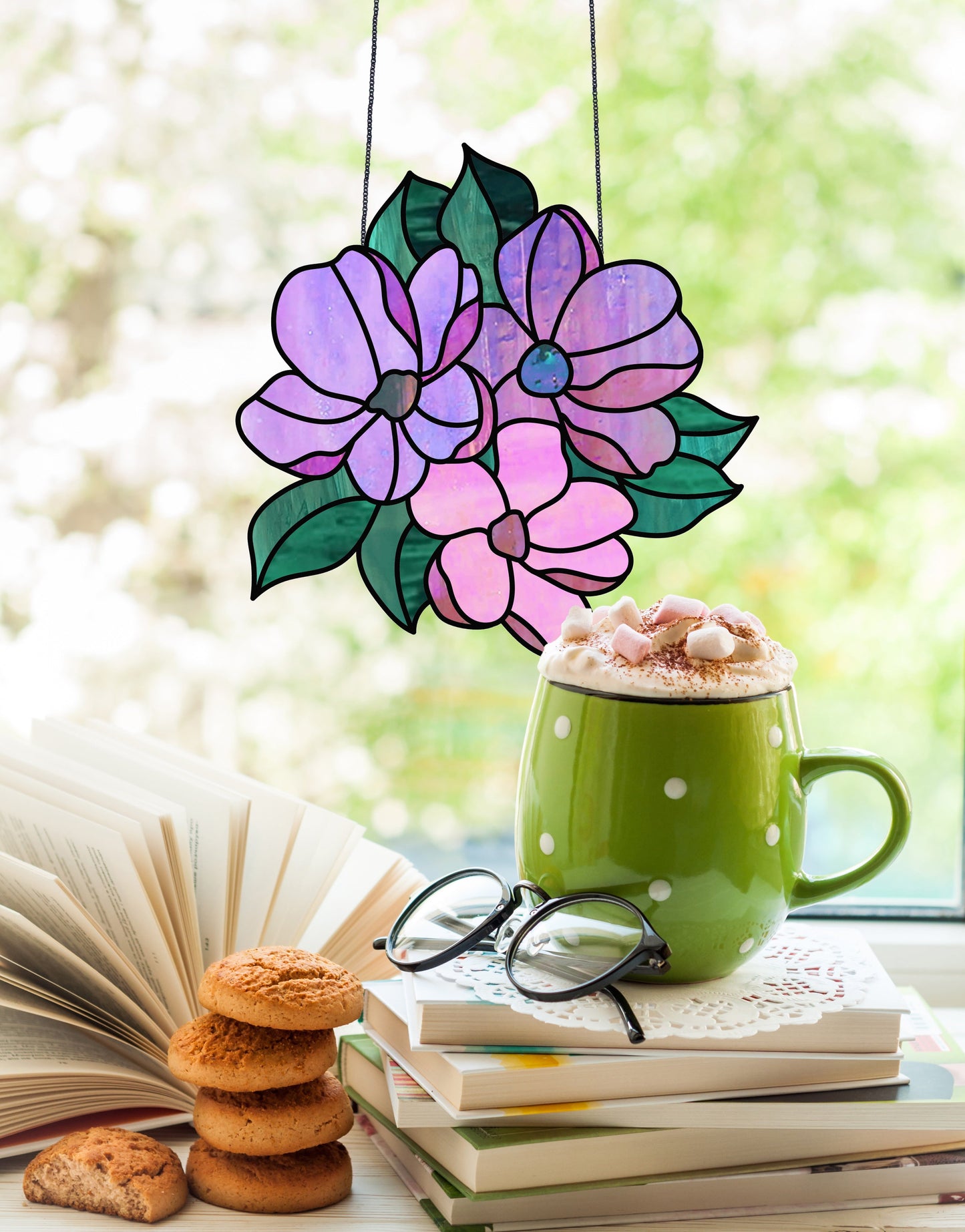 A green polka dot mug of hot chocolate and marshmallows rests on open books beside reading glasses. The Bundle of Flowers stained glass pattern hangs in the window. Nearby, fresh cookies are stacked, with blurred greenery in the backdrop.