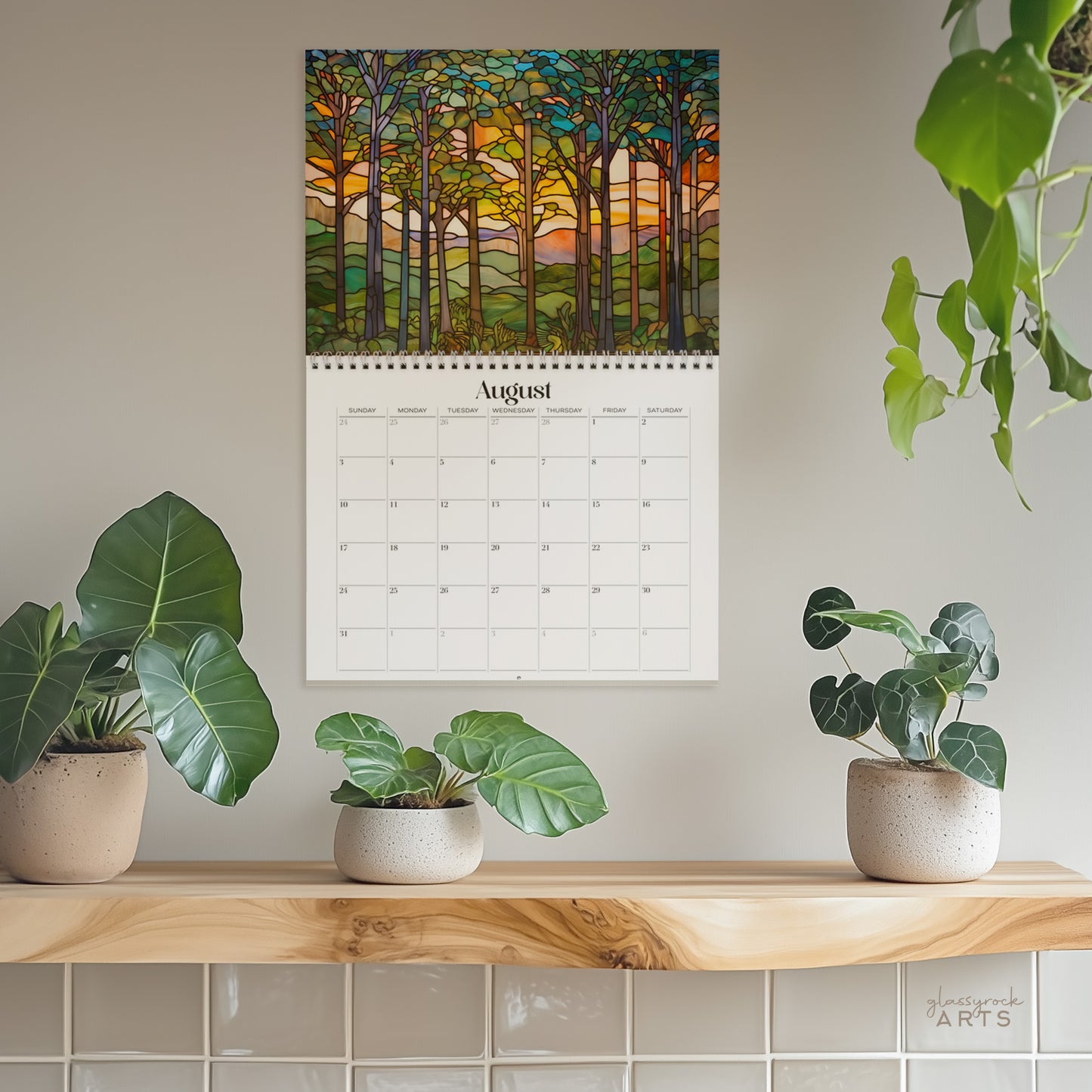 The forests calendar, a 2025 monthly wall calendar with forest-themed original artwork, hanging on a wall surrounded by plants. The August pages shows a serene forest sunset.