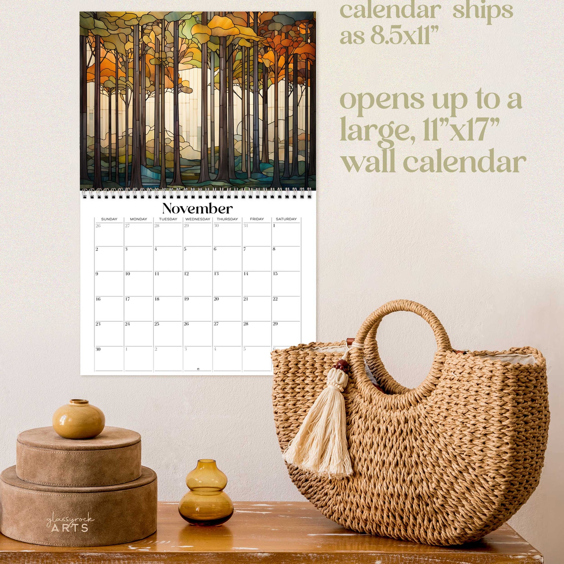 The forests calendar, a 2025 monthly wall calendar with forest-themed original artwork, on a wall above a wood table with a wicker purse. The calendar is open to November, showing a modern muted color forest.