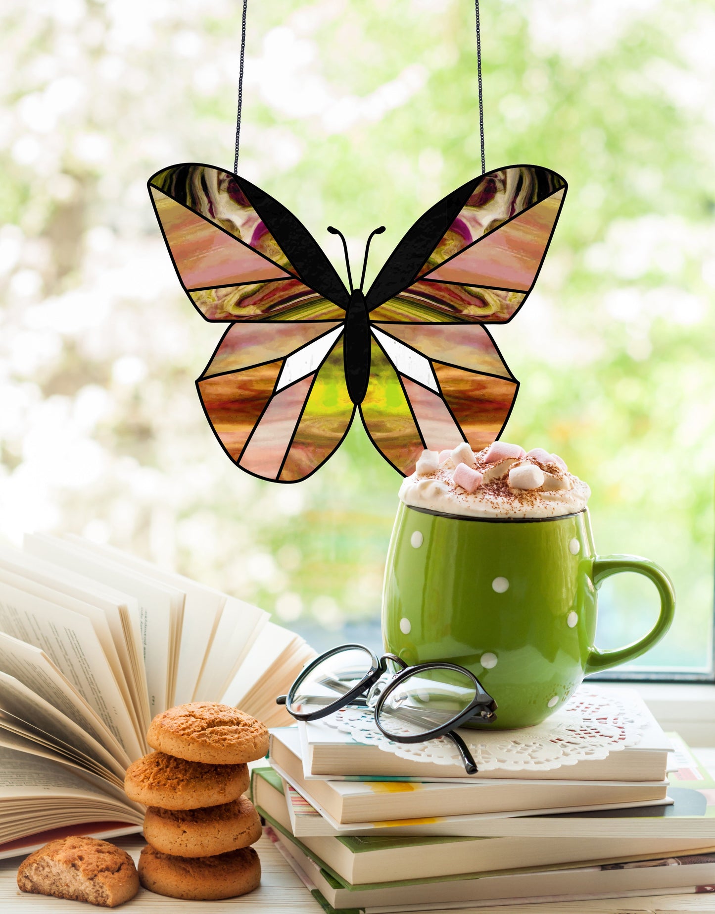 A Beginner Butterfly Stained Glass Pattern hangs in the window. On the sill, an open book is next to a green polka dot mug of hot chocolate with marshmallows, a stack of cookies, and glasses on books. The background is softly blurred.