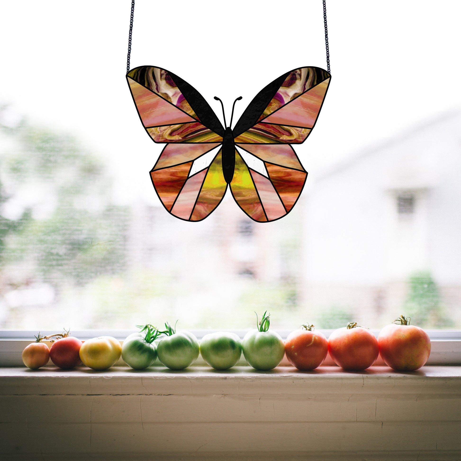 A Beginner Butterfly Stained Glass Pattern hangs in the window above a row of tomatoes, varying from green to red on the sill. A blurred garden is visible outside, ideal for those interested in digital pattern downloads or starting stained glass projects.