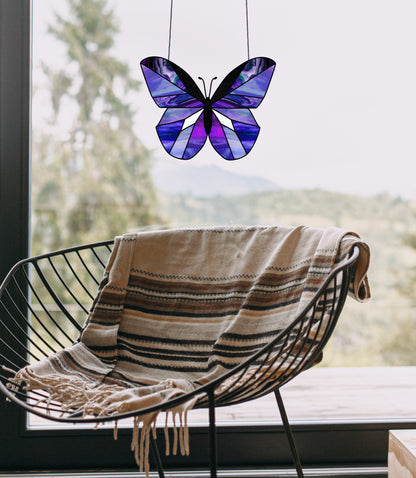 A black wire chair with a striped blanket sits by large windows. In front, the Beginner Butterfly Stained Glass Pattern hangs beautifully, offering a blurred view of trees and hills.