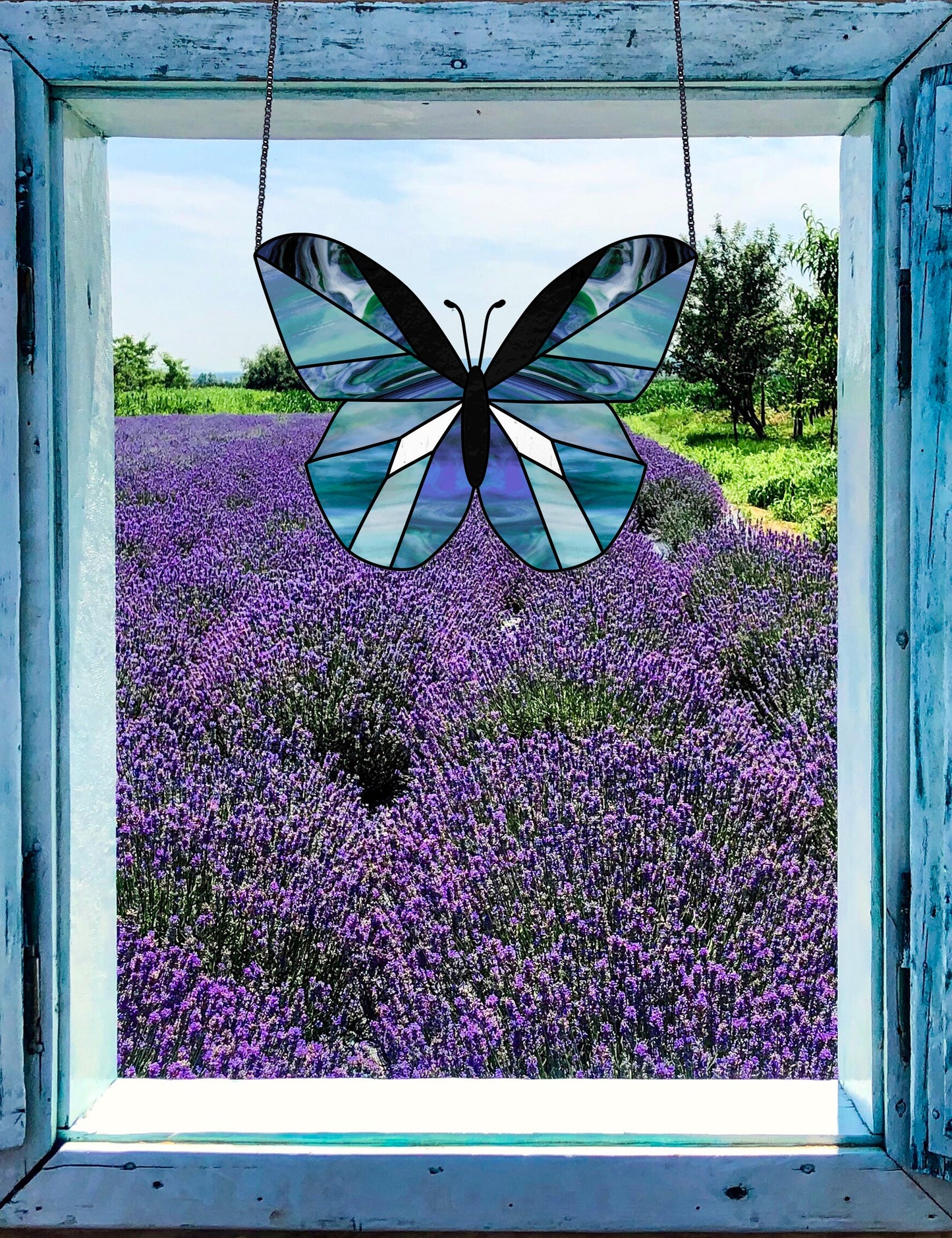 The Beginner Butterfly Stained Glass Pattern elegantly captures a butterfly framed by a weathered blue sill, hanging in a window overlooking lavender fields under a clear sky. Its detailed design mirrors the artistry in digital pattern downloads.