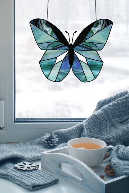 A stained glass butterfly from the Beginner Butterfly Stained Glass Pattern hangs by a window in blues and greens. Below, a wooden tray holds tea and pinecones, while a cozy gray sweater drapes nearby, all set against a snowy landscape.