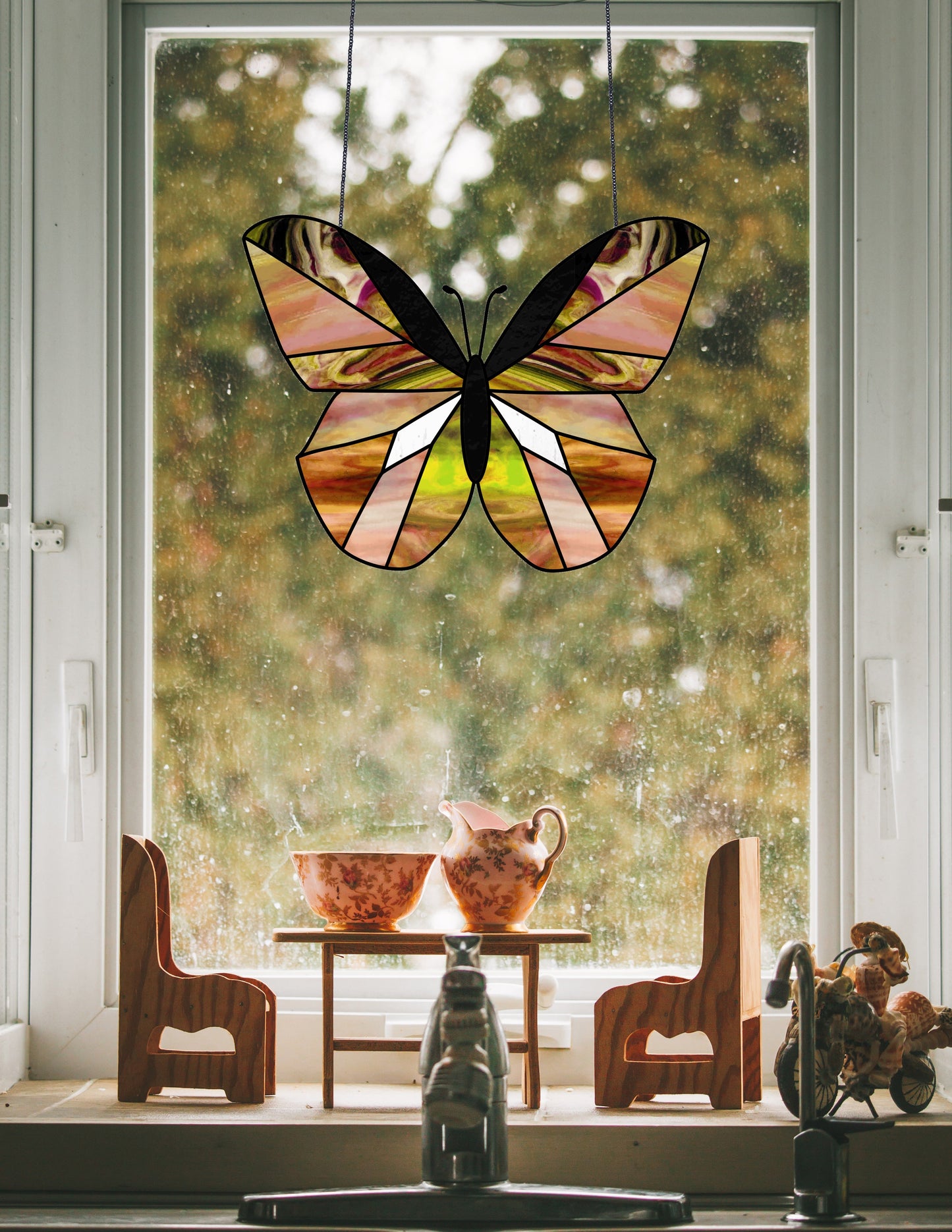 A Beginner Butterfly Stained Glass Pattern decorates a window above a wooden table for two, adorned with a teapot and floral bowl, against the blurred backdrop of trees, enhancing the scenes charming simplicity.