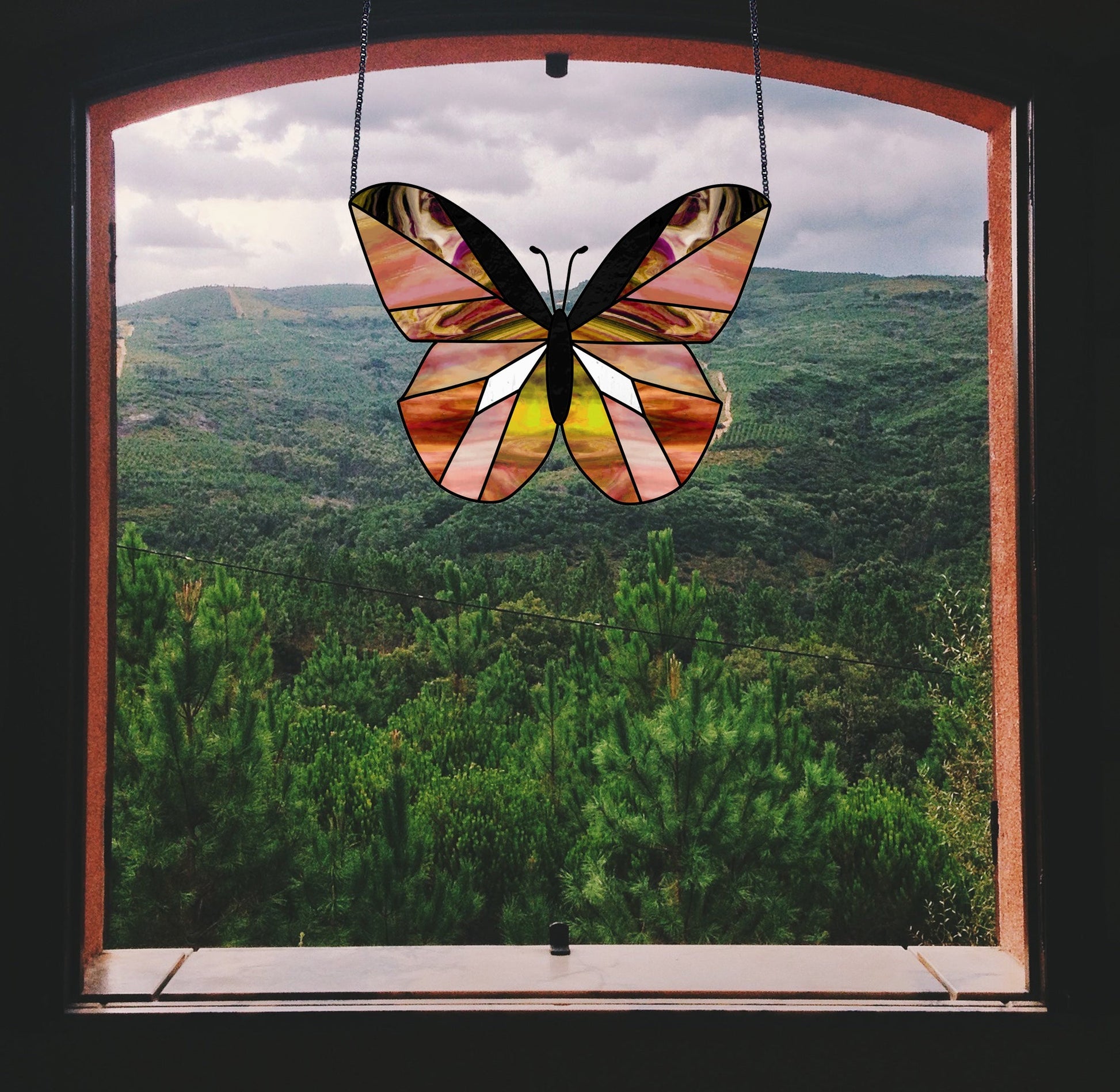 The Beginner Butterfly Stained Glass Pattern offers a vibrant mix of orange, purple, and yellow. It hangs elegantly in the center of an open window, contrasting beautifully against the lush green hillside under a cloudy sky.