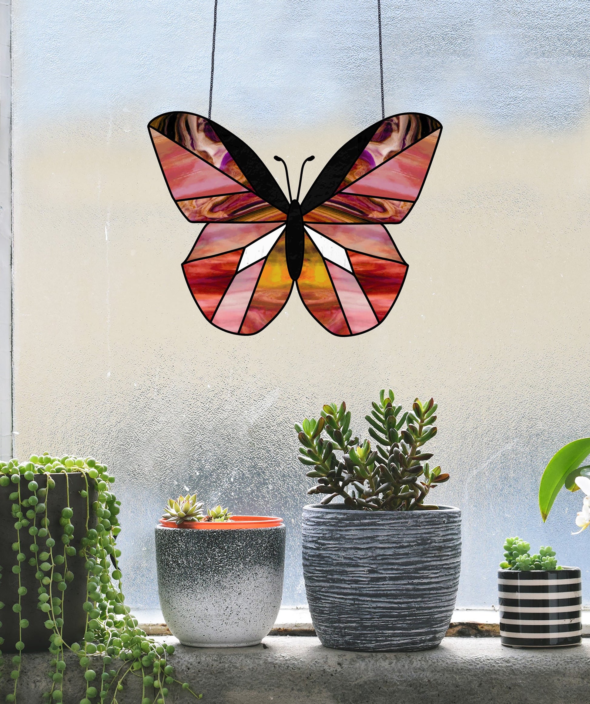 A stained glass butterfly, made using the Beginner Butterfly Stained Glass Pattern, hangs before a frosted window. On the sill below sit three potted plants: a trailing succulent, a gray speckled pot with a red rim, and a striped pot with blooms.