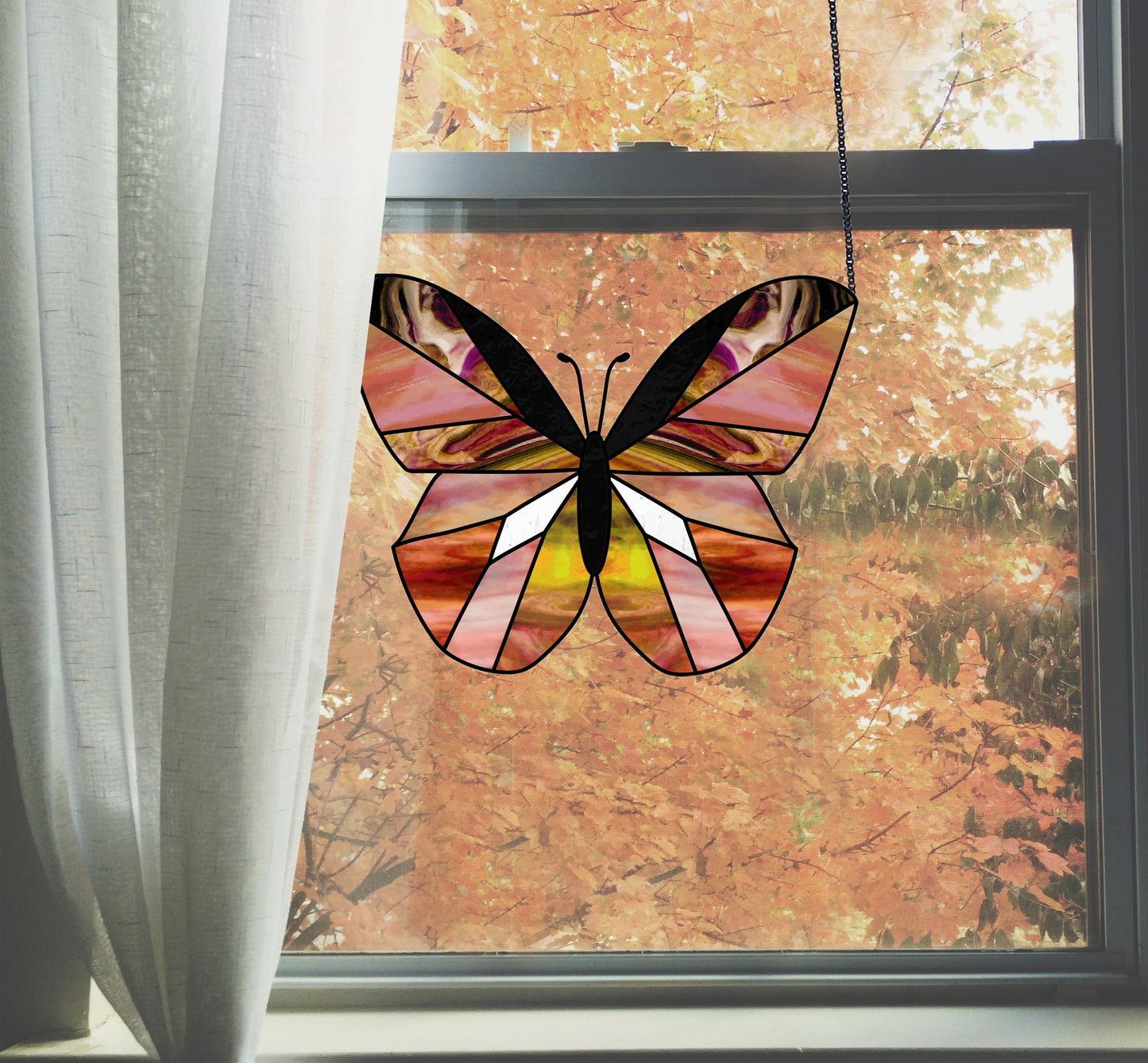 A butterfly, crafted from the Beginner Butterfly Stained Glass Pattern, hangs in front of a window with white curtains. Outside, an autumn scene unfolds with tree leaves in shades of orange and brown.