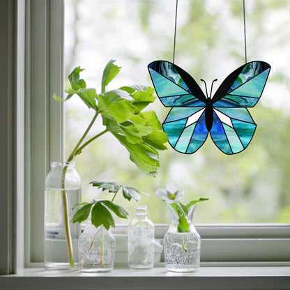 A Beginner Butterfly Stained Glass Pattern in blue shades hangs in a window, accompanied by three glass bottles with leafy plants and a white flower for a peaceful, airy look.