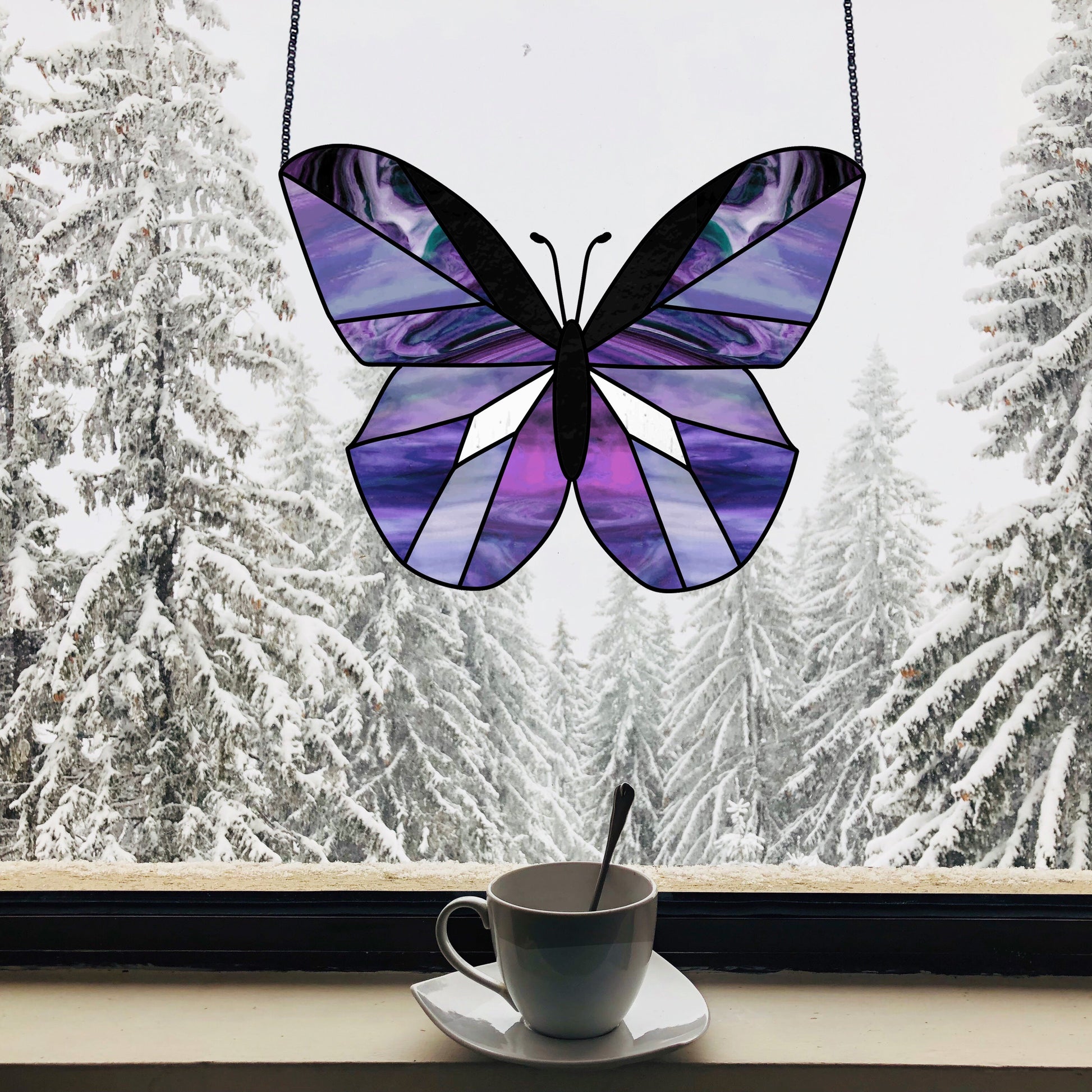 The Beginner Butterfly Stained Glass Pattern, casting purple hues through the window, creates colorful shadows on a snowy wintry scene. A white cup with a spoon sits on the saucer by the windowsill, completing this peaceful tableau—ideal for novice stained glass crafters.