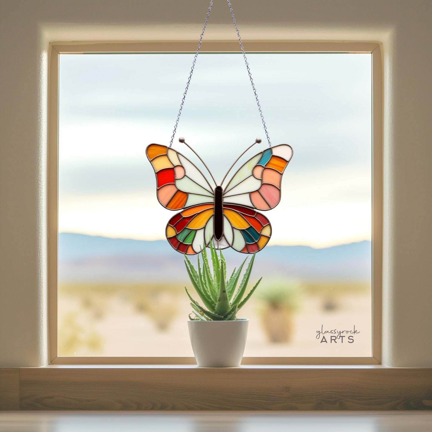 Stained glass butterfly art hanging in a window, with a potted succulent on the windowsill and a desert landscape visible outside.