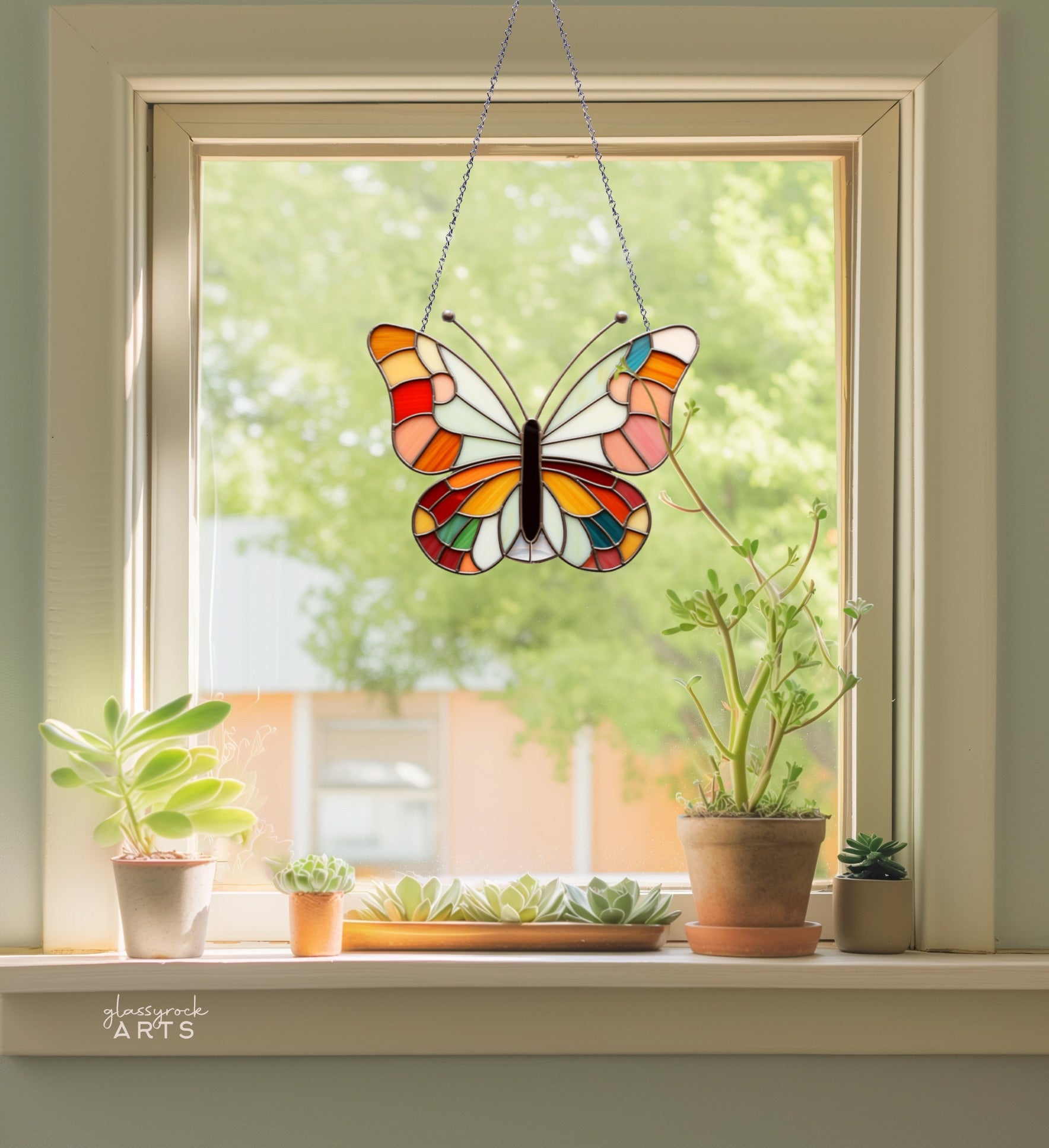 A colorful stained glass butterfly hangs in a window, surrounded by potted succulents on the windowsill with a blurred view of greenery outside.