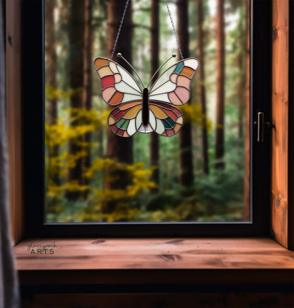 A stained glass butterfly sun catcher hangs by a chain on a wooden-framed window, with a blurred forest scene in the background.