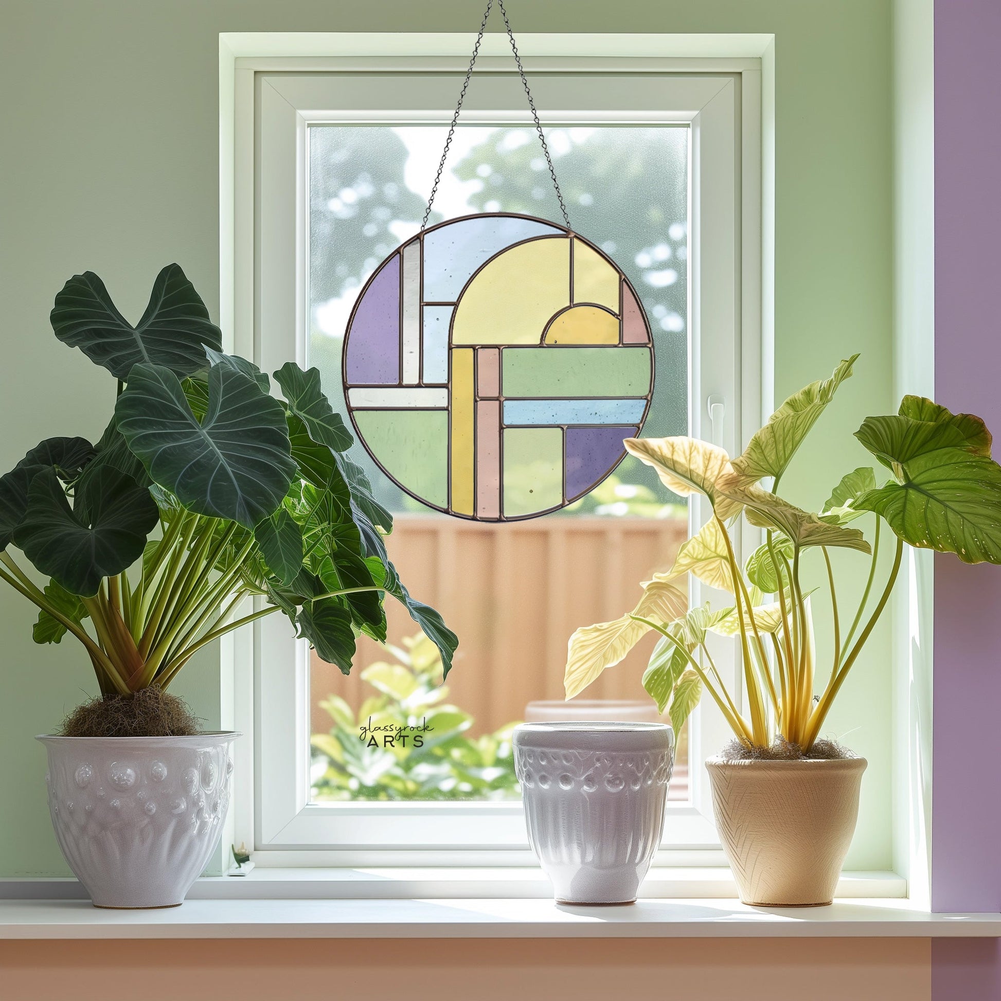 A round, multicolored stained glass piece hangs in front of a window. Two potted plants sit on the windowsill, one with large green leaves and the other with yellowish leaves.