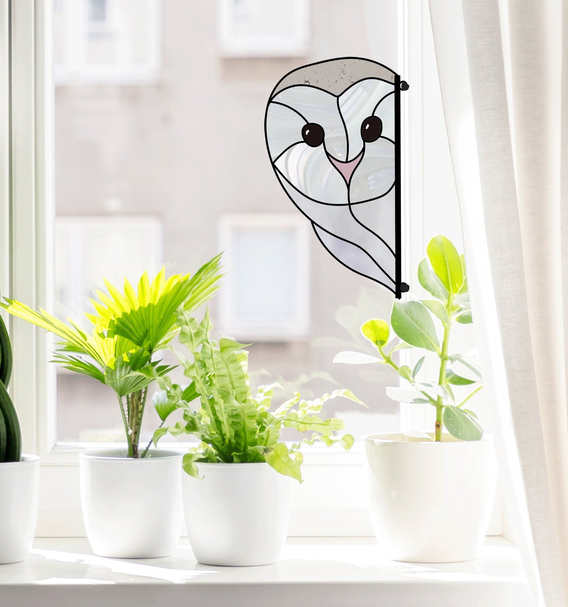 Three potted plants sit on a windowsill with the Snowy Owl Stained Glass Pattern beside them. The open window, framed by sheer white curtains, allows bright natural light to highlight the art piece, which can be replicated with a digital pattern download for crafting enthusiasts.