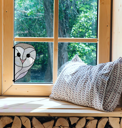 The Snowy Owl Stained Glass Pattern adds charm to a wooden window, available as a digital download. Sunlight illuminates a cozy nook with a patterned pillow, while outside, green trees and neatly stacked logs enhance the peaceful setting.