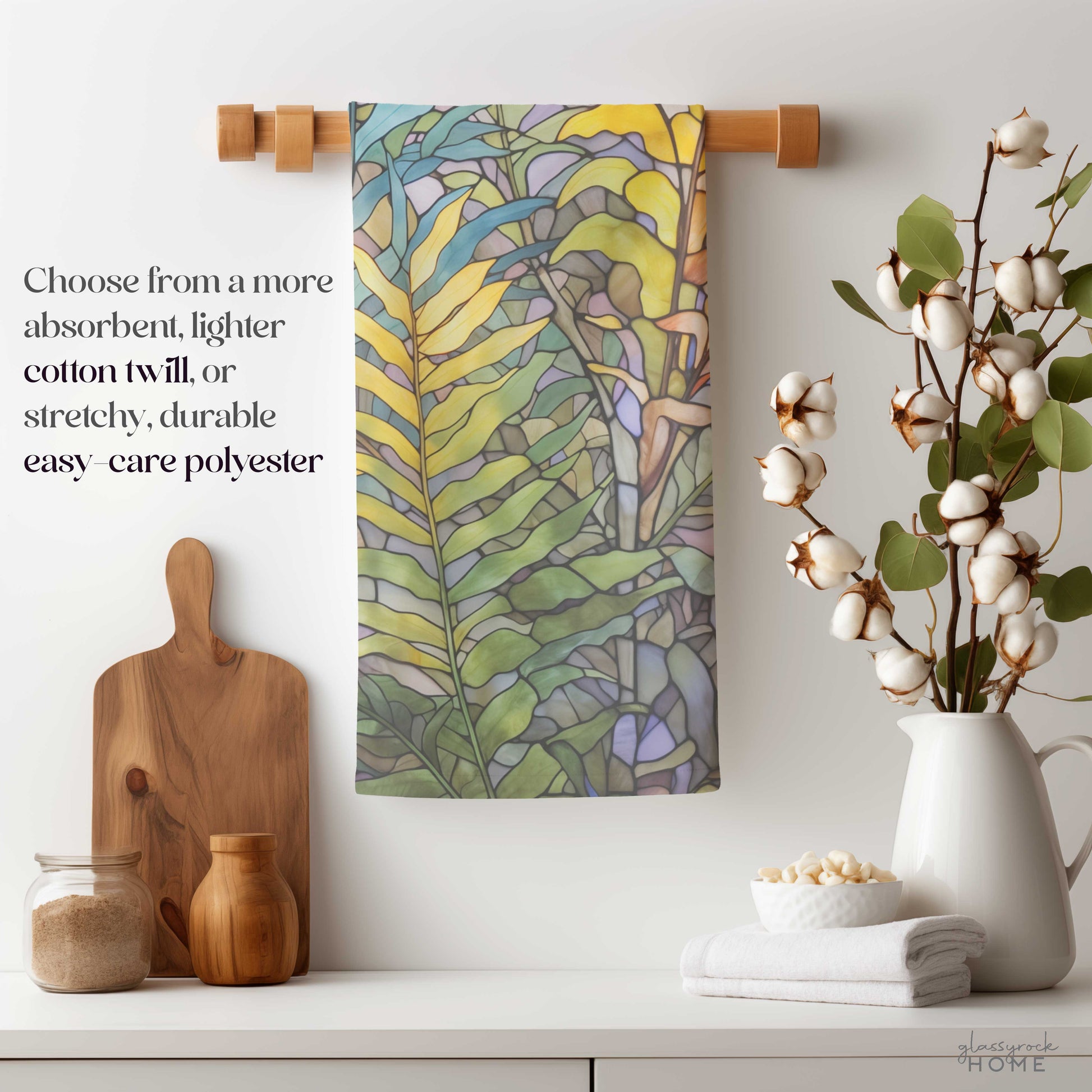 A Stained Glass Pastel Ferns Dish Towel elegantly hangs on a wooden rod above the kitchen counter, complemented by jars, a cutting board, cotton stems in a vase, and a neat stack of white towels. The text highlights fabric choices such as easy-care soft polyester and natural cotton twill.