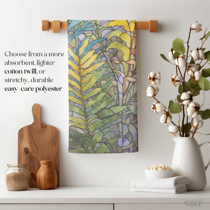 A Stained Glass Pastel Ferns Dish Towel elegantly hangs on a wooden rod above the kitchen counter, complemented by jars, a cutting board, cotton stems in a vase, and a neat stack of white towels. The text highlights fabric choices such as easy-care soft polyester and natural cotton twill.