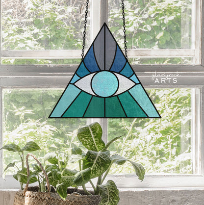 The Boho Eye Triangle Stained Glass Pattern, featuring a digital blue and green eye design, hangs by a chain in front of a bright window. Below, a windowsill holds a potted plant with broad variegated leaves.