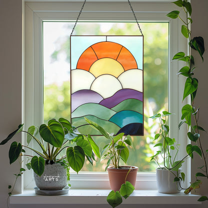 A fun curvilinear stained glass piece with layered mountains, clouds, sun and sky. It is hanging in a window surrounded by plants.