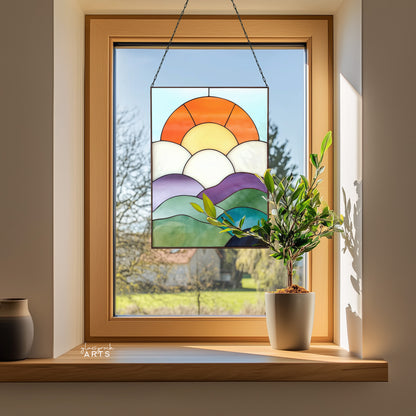 A fun curvilinear stained glass piece with layered mountains, clouds, sun and sky. It is hanging in a window surrounded by a small olive tree plant and a vase.