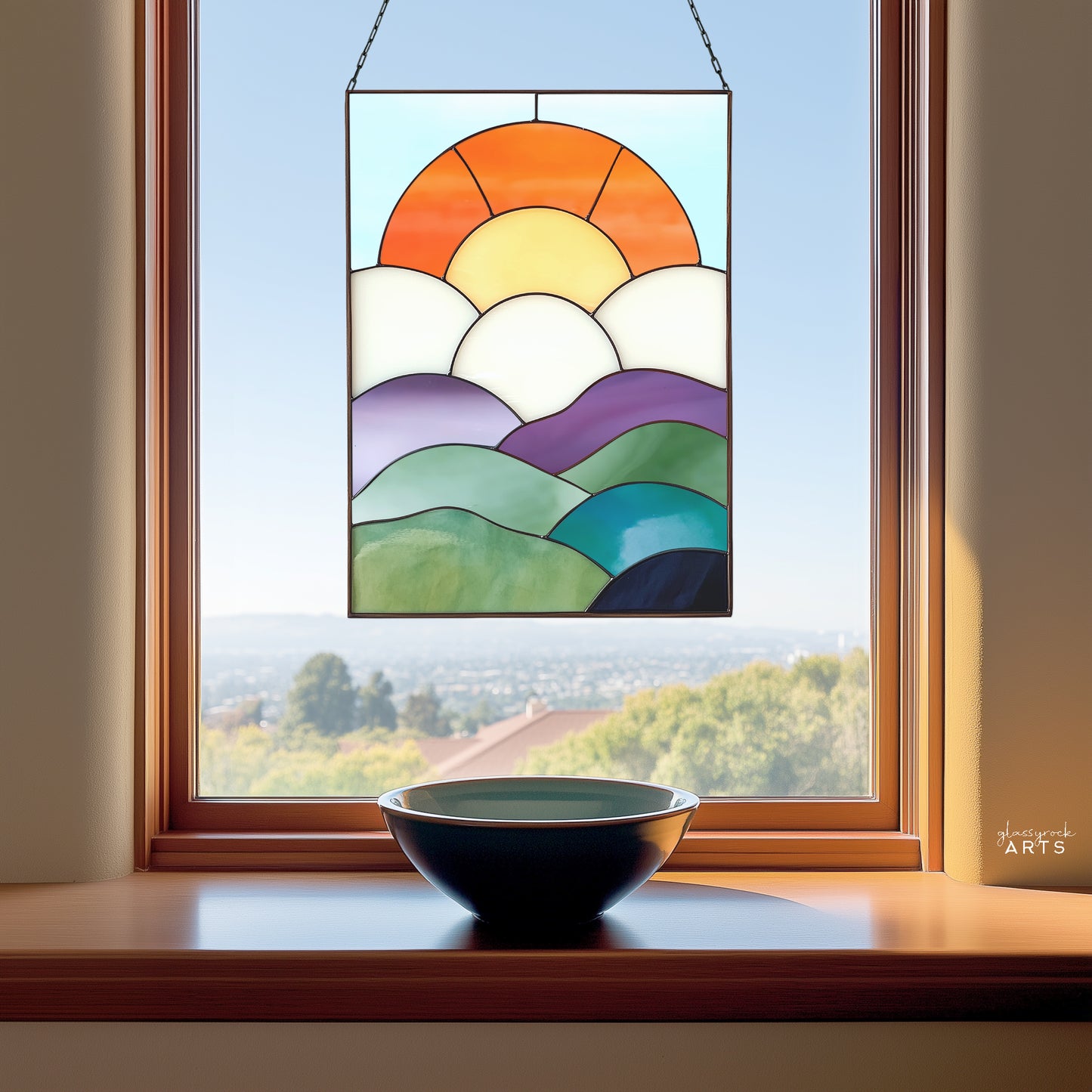 A fun curvilinear stained glass piece with layered mountains, clouds, sun and sky. It is hanging in a window above a ceramic bowl.