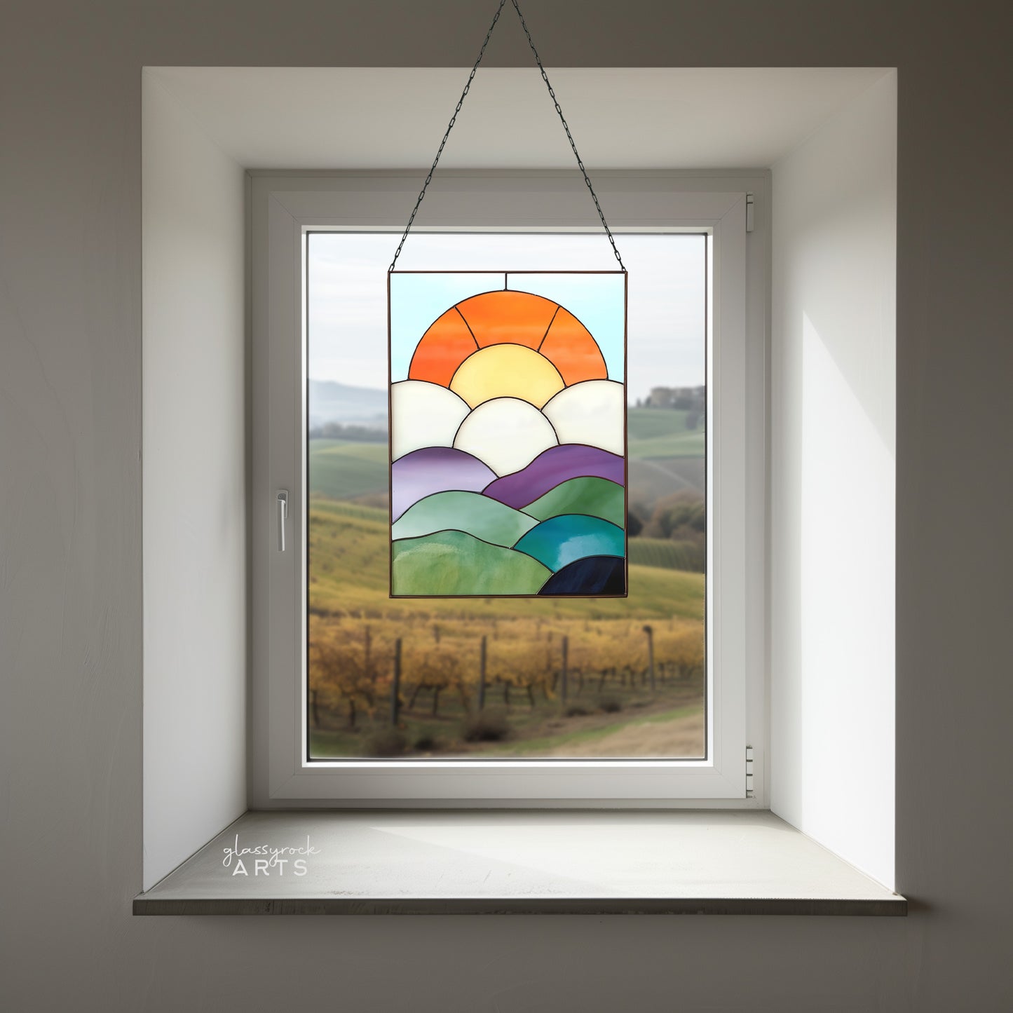 A fun curvilinear stained glass piece with layered mountains, clouds, sun and sky. It is hanging in a white window.