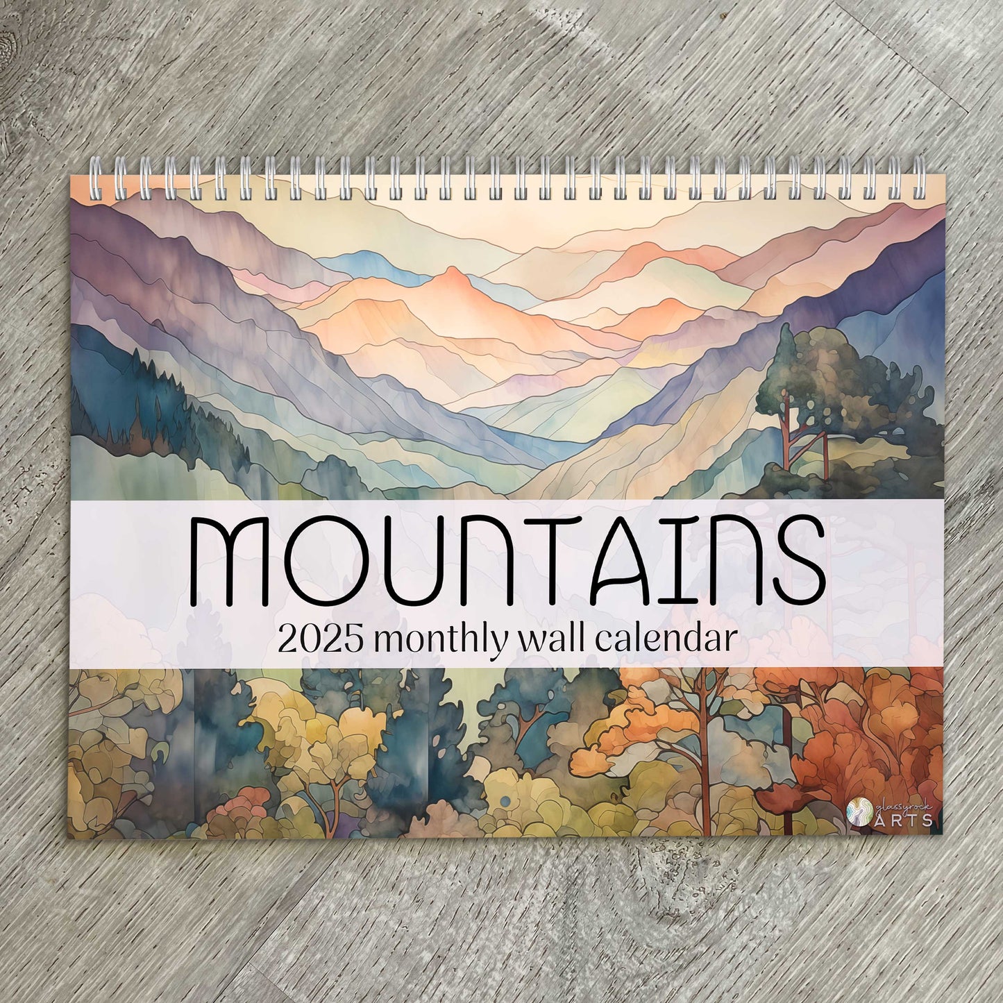 The mountains calendar, a 2025 monthly wall calendar with mountains-themed original artwork, on a wood table surface. 