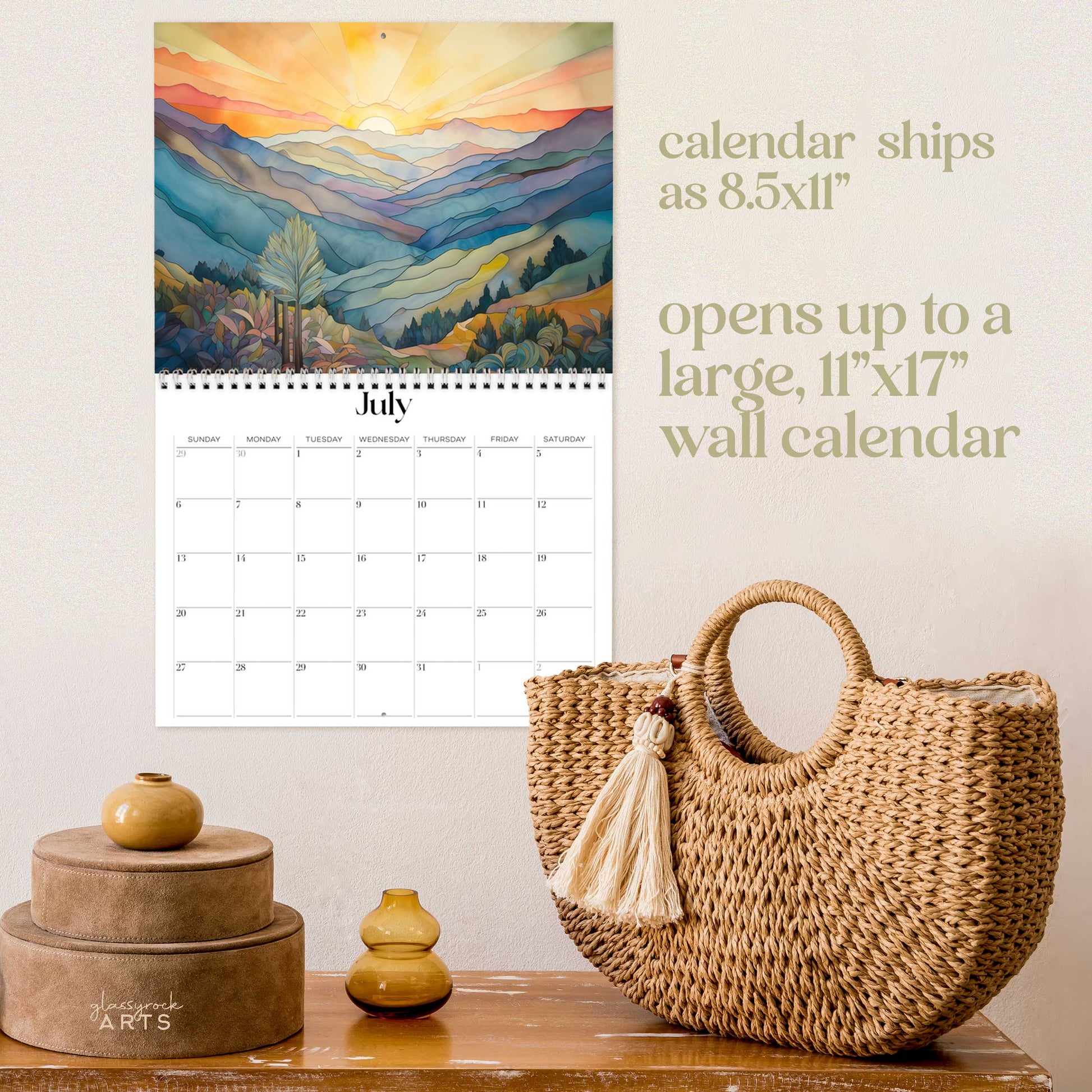 The mountains calendar, a 2025 monthly wall calendar with mountain-themed original artwork, on a wall above a wood table with a wicker purse. The calendar is open to July, showing a muted rainbow-colored mountain landscape at sunset.