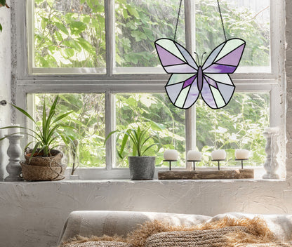 A cozy room features a large window with a vibrant Beginner Butterfly Stained Glass Pattern hanging. Below are potted plants and candles on a wooden tray. A sofa with textured pillows sits in front of the window, where rich greenery peeks through outside.