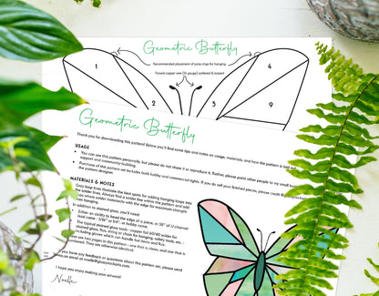 A Beginner Butterfly Stained Glass Pattern rests on wood surrounded by green ferns. It features geometric sections and numbered instructions for crafting, similar to a digital download for easy guidance.