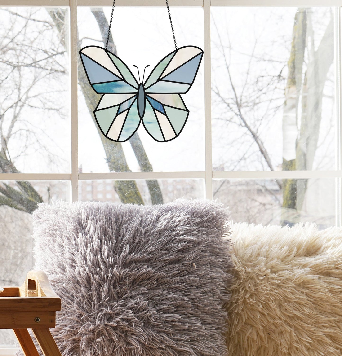 A Beginner Butterfly Stained Glass Pattern in blue and white hangs near two pillows, one gray and one cream, framing a view of bare trees. Ideal for art lovers with a downloadable digital pattern.