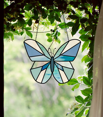 A stained glass butterfly, created using the Beginner Butterfly Stained Glass Pattern and featuring blue and white hues, hangs among green foliage, its beauty resonating against the blurred leafy backdrop beyond the window.