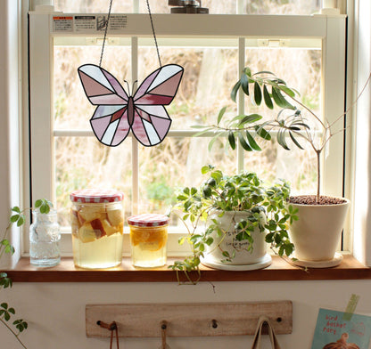 A window sill adorned with a stained glass butterfly glows in sunlight, illuminating potted plants, jars with checkered lids, and a pitcher. Perfect for those inspired to start a beginner stained glass project or download the Beginner Butterfly Stained Glass Pattern for an artistic touch.