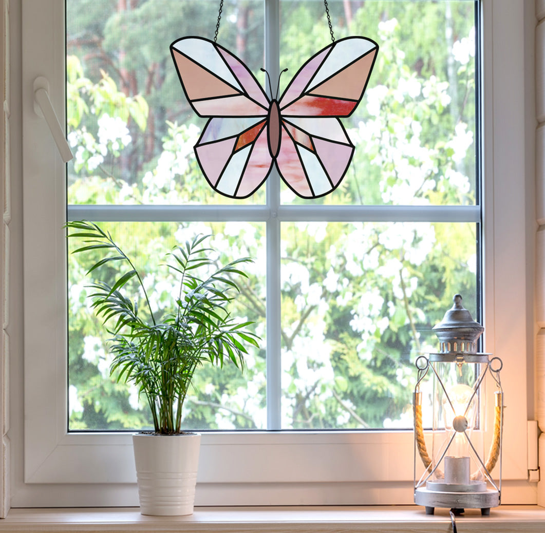 A window displays the Beginner Butterfly Stained Glass Pattern hanging at its center, ideal for digital pattern seekers. A potted plant and lantern grace the sill, framed by lush trees and white flowers outside.