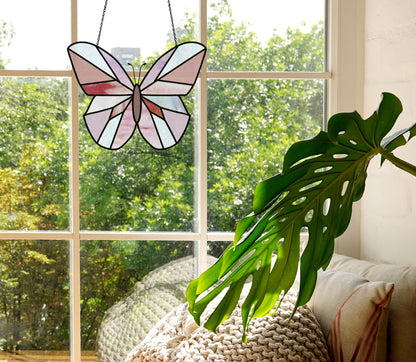 A Beginner Butterfly Stained Glass Pattern suncatcher in pink and purple hangs in a sunny window. Below, a cozy beige blanket and pillow lie on a sofa, while a large monstera leaf in the foreground adds greenery.