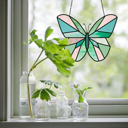 A Beginner Butterfly Stained Glass Pattern features a green and peach stained glass butterfly hanging in a window with glass bottles holding leaves below, set against blurred greenery outside. Ideal as a stained glass pattern or digital download.