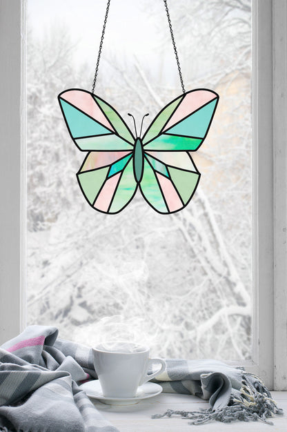 A Beginner Butterfly Stained Glass Pattern with pastel colors adorns a window, revealing a snowy view outside, while inside, a steaming coffee cup rests on the sill under a gray and pink blanket.