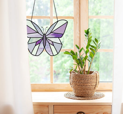 A Beginner Butterfly Stained Glass Pattern in purple and white hues adorns a wooden-framed window. Nearby, a green potted plant sits on the wooden surface, while white curtains frame the vibrant leafy view outside.