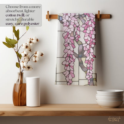 A Stained Glass Pink Wisteria Dish Towel hangs on a wooden peg. Nearby are a vase with cotton stems, paper towels, and stacked white bowls. A sign highlights its natural cotton twill material option.