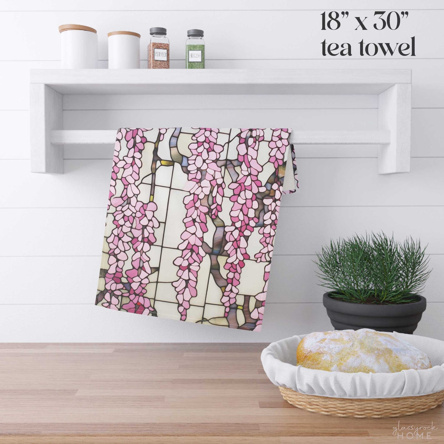 The Stained Glass Pink Wisteria Dish Towel, crafted from natural cotton twill and measuring 18 x 30, hangs on a white wall shelf adorned with small jars, while beneath it sits a bread loaf next to a potted plant.