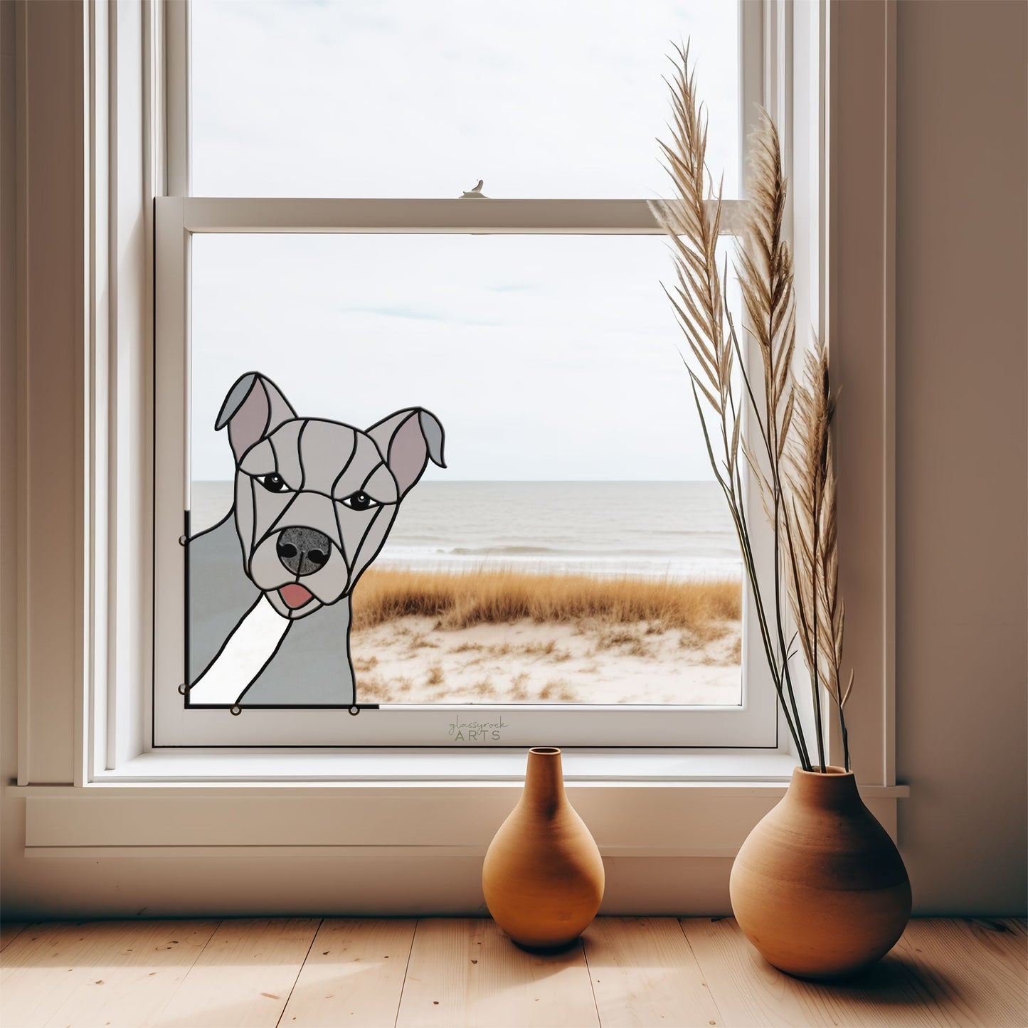 A window reveals a coastal view with sand dunes beneath a cloudy sky, adorned with an American Pitbull Terrier Dog Stained Glass Pattern. Two ceramic vases with dried grasses rest on the wooden floor, adding charm to this seaside sanctuary.