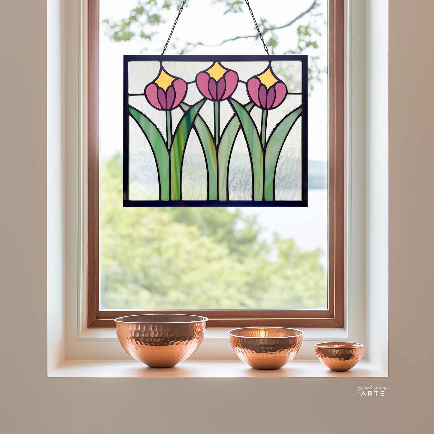 A beautiful rectangular stained glass flower panel, prairie style with three pink and yellow tulips and green leaves with clear glass background, hanging in a window with a pastoral lake background and three copper bowls on the windowsill.