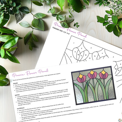 Two pages of the Prairie Flowers Panel stained glass pattern on a pale grey wood table with plants.