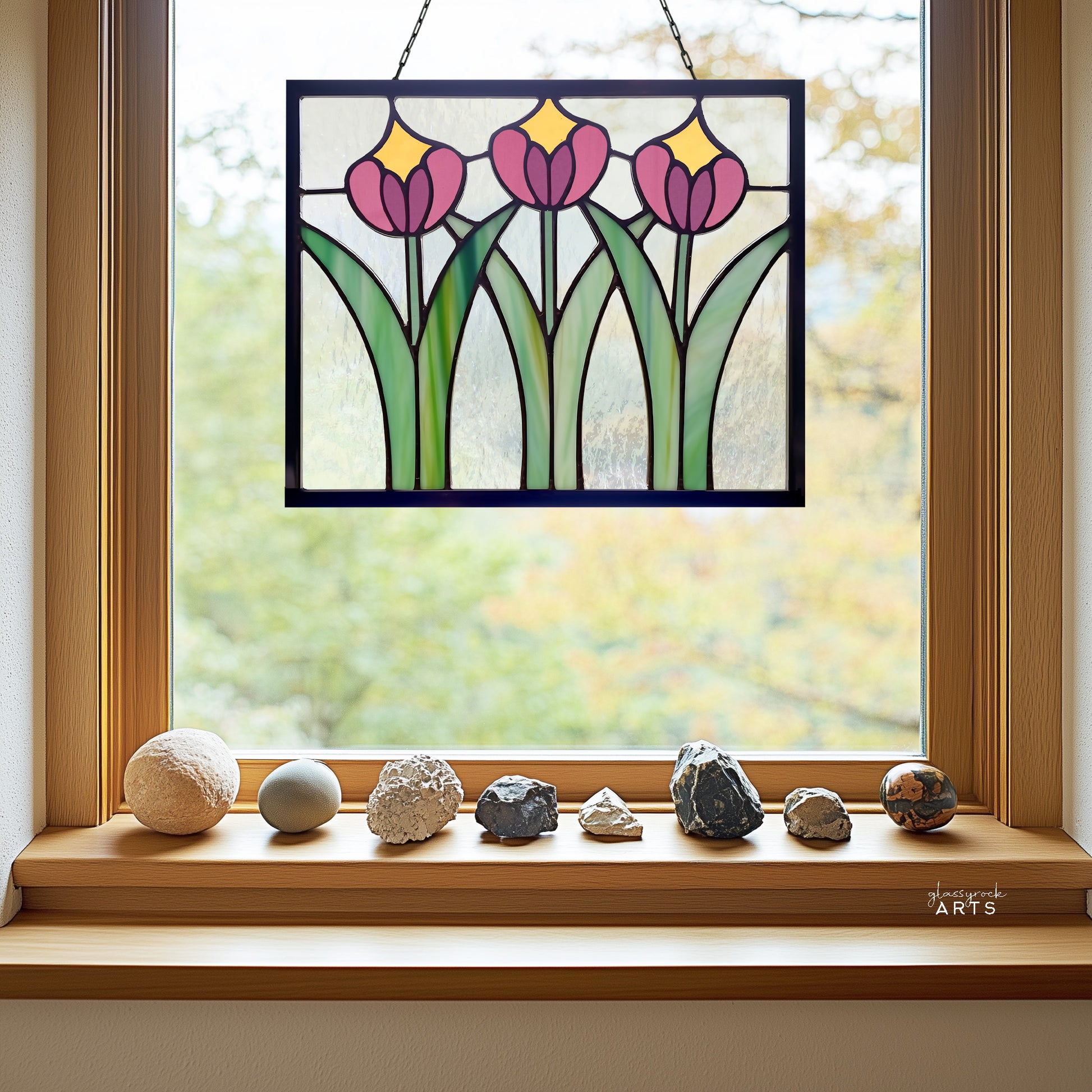 A beautiful rectangular stained glass flower panel, prairie style with three pink and yellow tulips and green leaves with clear glass background, hanging in a window with a green trees background and a rock collection on the windowsill.