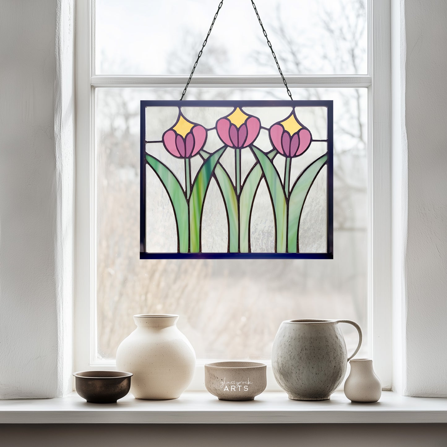 A beautiful rectangular stained glass flower panel, prairie style with three pink and yellow tulips and green leaves with clear glass background, hanging in a window with a winter trees background and a ceramic bowl and pitcher collection on the windowsill. 