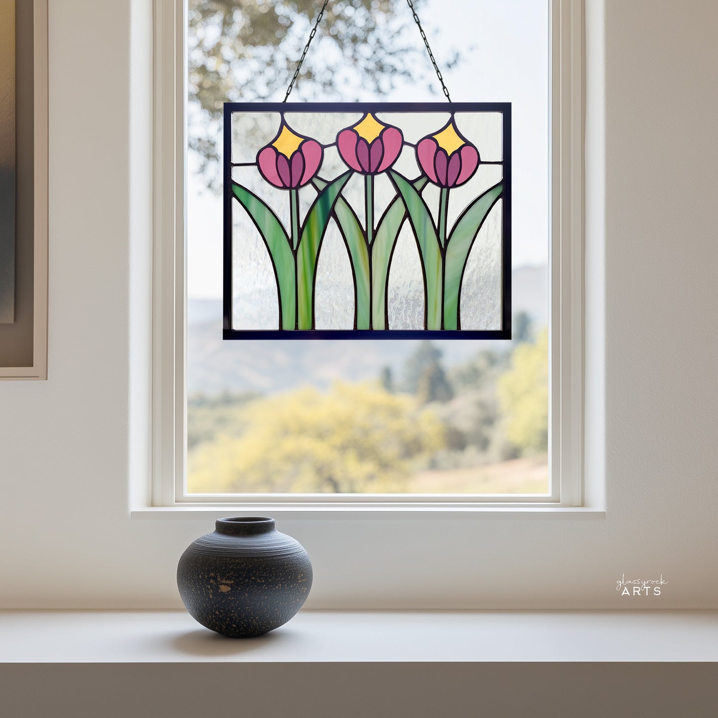 A beautiful rectangular stained glass flower panel, prairie style with three pink and yellow tulips and green leaves with clear glass background, hanging in a window with a fall trees background and a small ceramic vase on the windowsill.