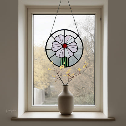 A round stained glass panel with a pink flower and red center, hanging in a window with a winter trees background, with a vase on the windowsill. 